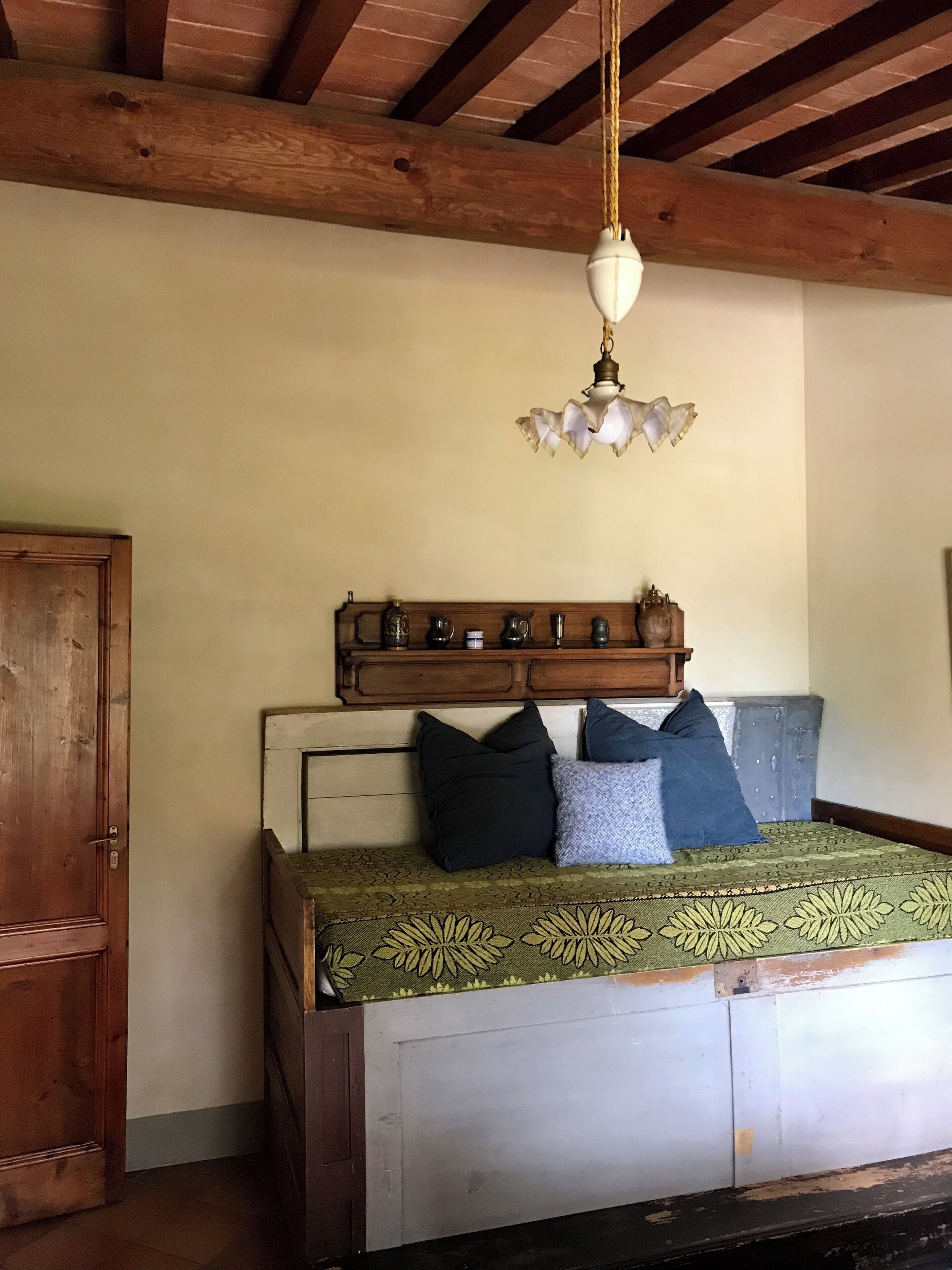 A bedroom with a wooden bed frame, a green patterned bedspread, and a wooden shelf above the bed holding various items.
