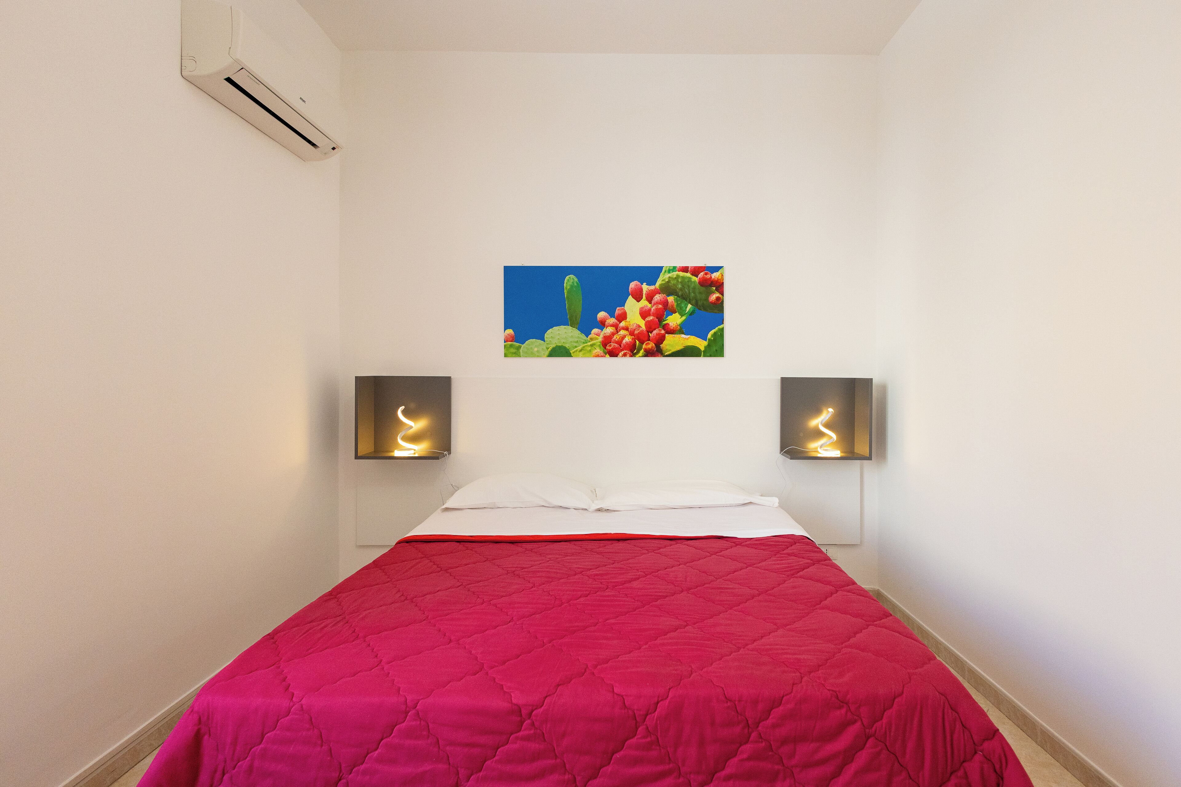A bedroom with a red quilted bedspread, a painting of a cactus and flowers on the wall, and two wall-mounted speakers.