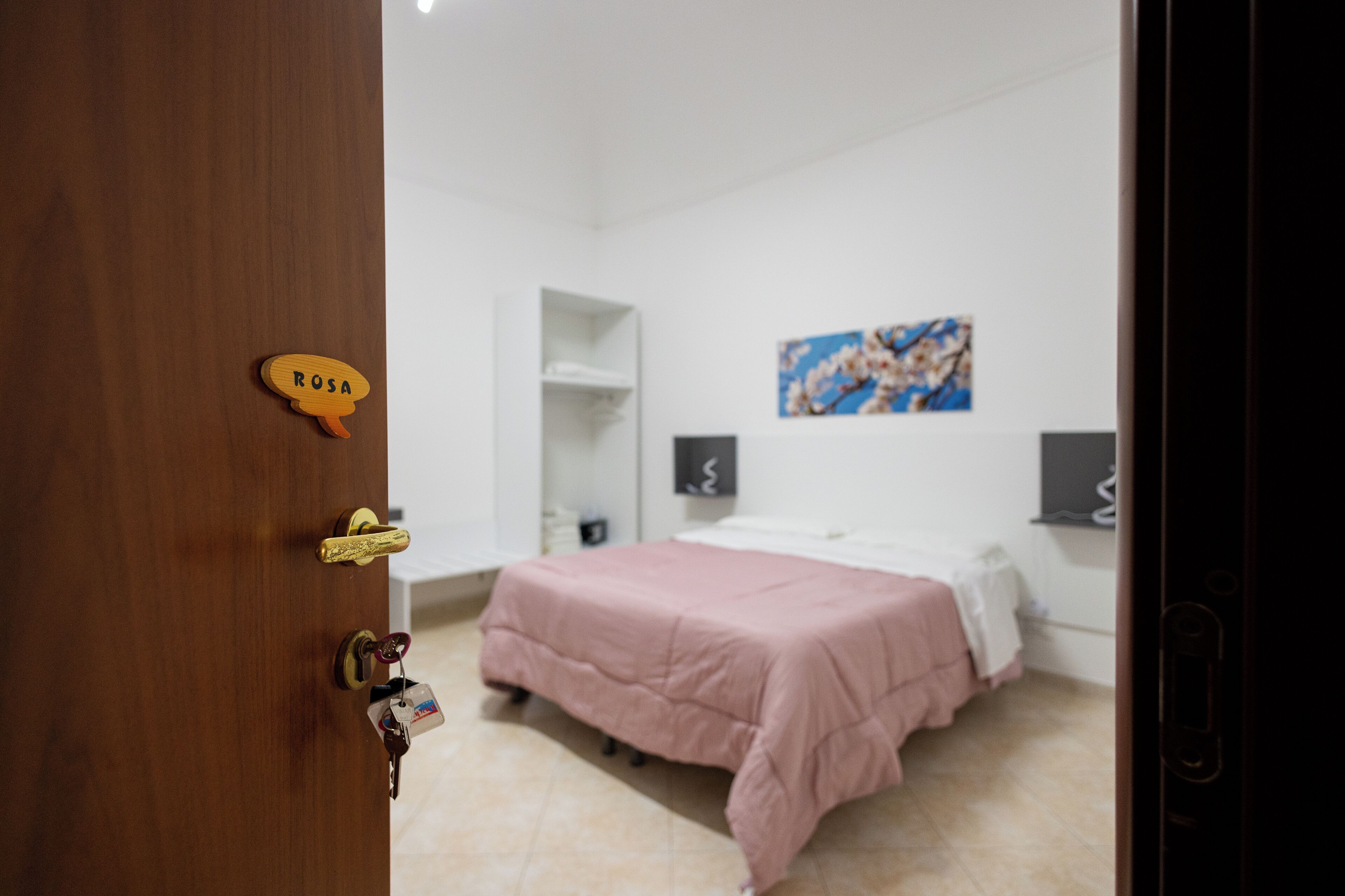 Un dormitorio con una cama, una puerta de madera con una placa, un estante blanco y un cuadro en la pared.