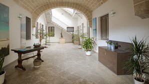 A spacious hallway with a high ceiling, tiled floor, and a staircase leading to an upper level. There are potted plants along the walls and a reception desk at the end of the hallway. - CasaTrapani (Trapani)
