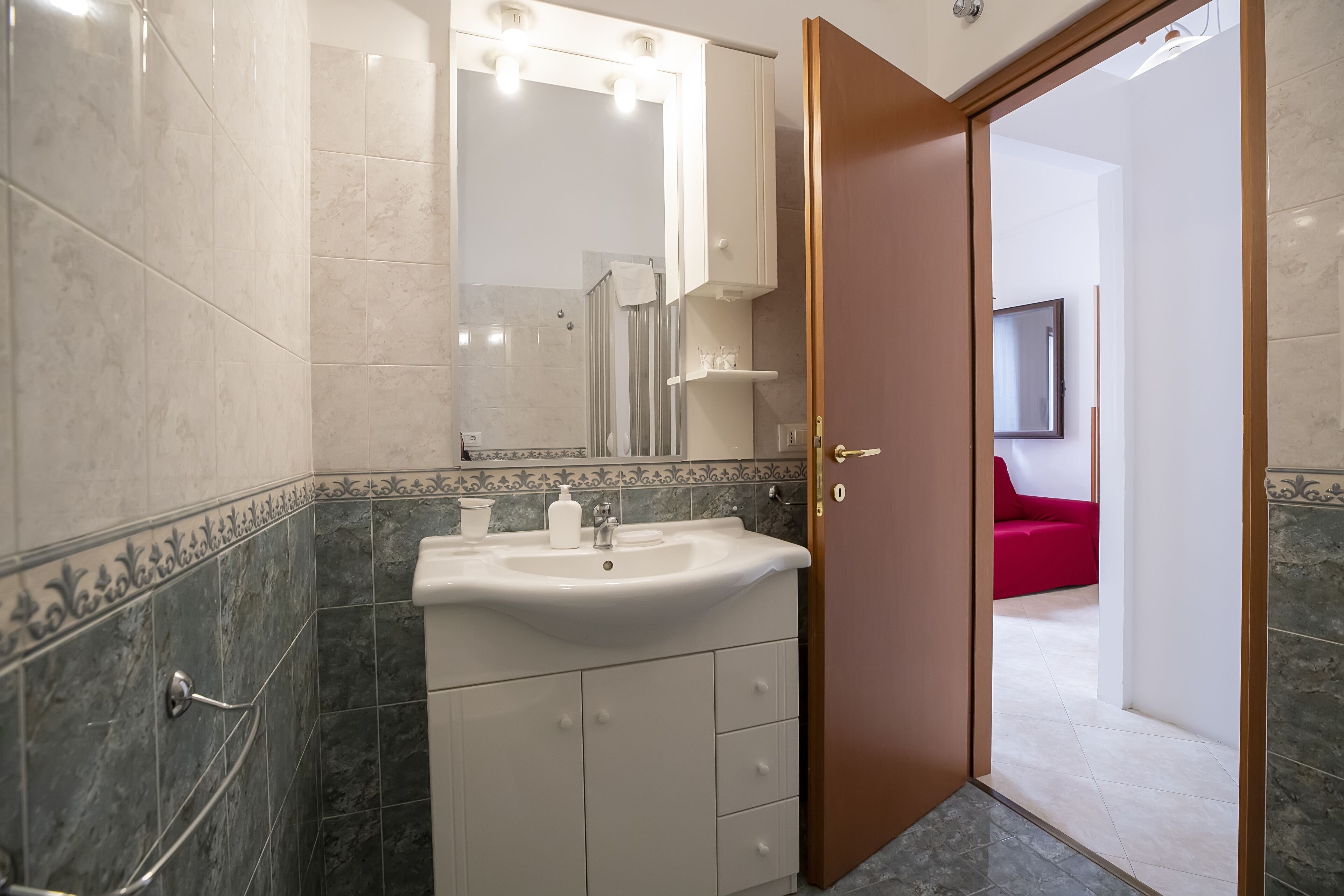 A bathroom with a sink, mirror, and shower. A door leads to a room with a red sofa.