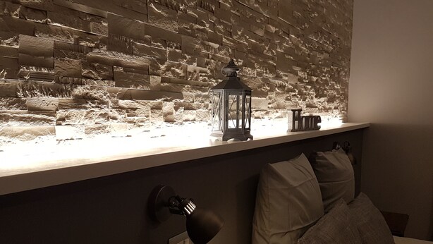 A room with a textured wall, a small table with a lantern, a bed with pillows, and a wall-mounted lamp. - La Locanda dell'Angelo (Rome)