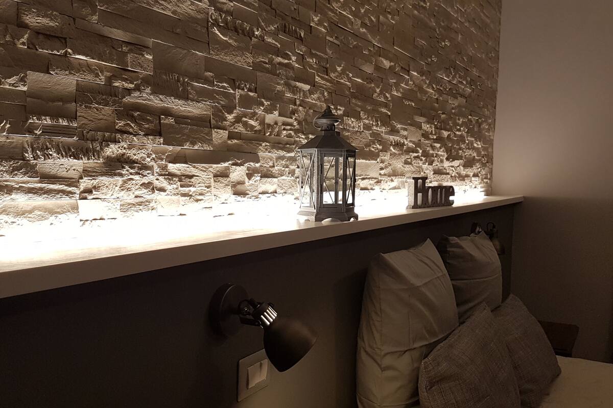 A room with a textured wall, a small table with a lantern, a bed with pillows, and a wall-mounted lamp.
