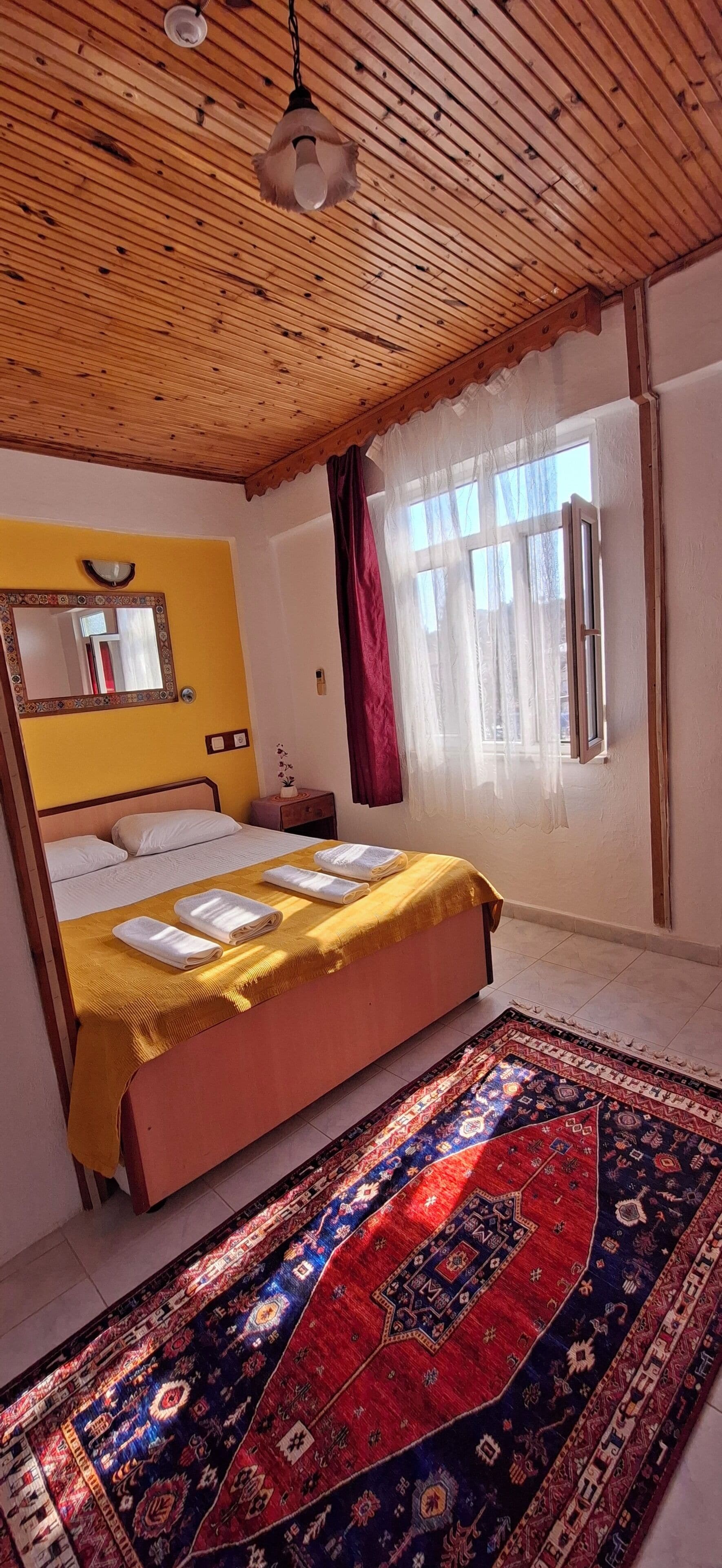 Ein Zimmer mit einem Bett, Holzboden, einem Fenster mit Vorhängen, einem Spiegel und einem gemusterten Teppich.