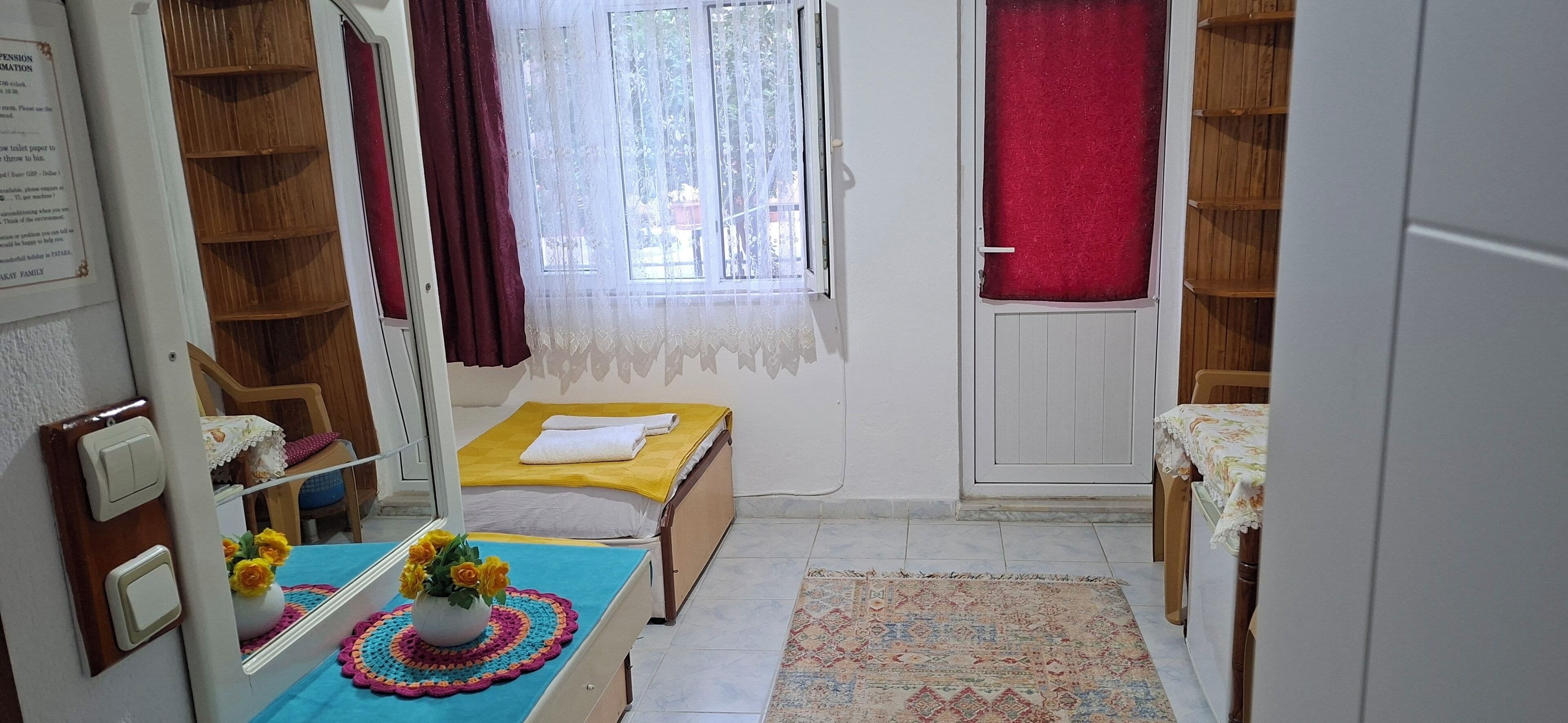 A compact room with a bed, a small table with a vase of flowers, a window with a sheer curtain, and a door with a red curtain.