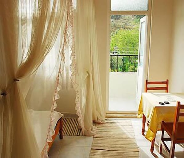 A room with a bed, a table, and a chair, a window with curtains, and a view of greenery outside.