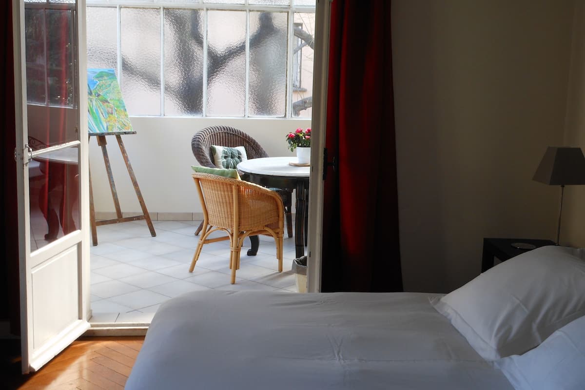 A room with a bed, a wicker chair, a small table with a vase of flowers, and a painting on an easel by a window.