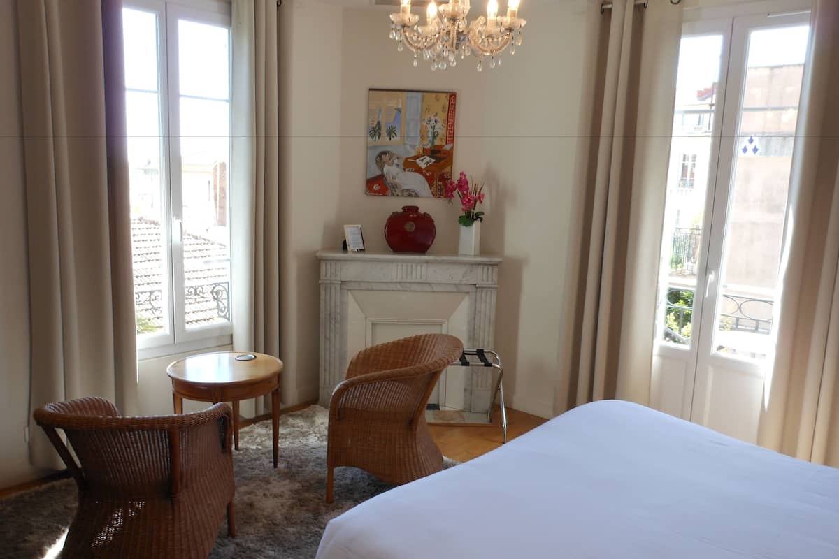 A hotel room with a large bed, two wicker chairs, a small round table, a fireplace, and a painting on the wall.