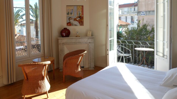 Un dormitorio con cama, una mesa de comedor, dos sillas de mimbre, chimenea y balcón con vistas a edificios y árboles.