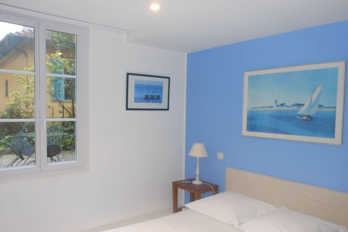 A bedroom with a bed, a nightstand with a lamp, a framed picture on the wall, and a window with a view of the outside.