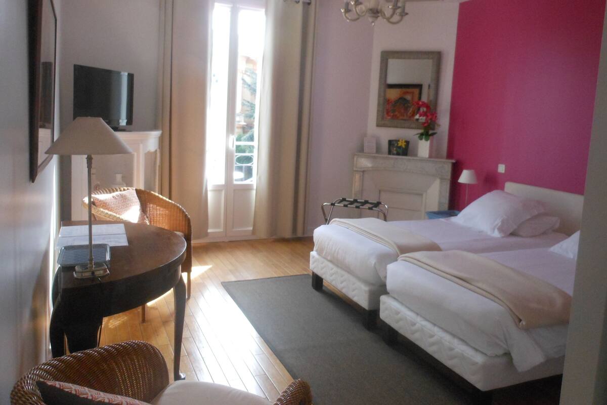 A hotel room with a bed, a desk, a chair, a small table, a mirror, and a window with curtains.
