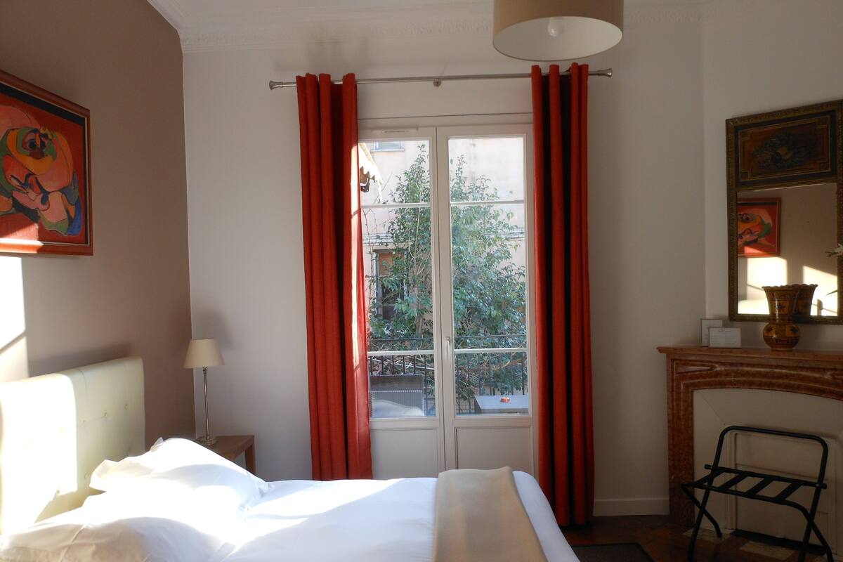 A hotel room with a bed, a chair, a fireplace, and a window with red curtains.