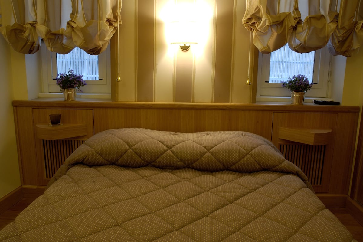 A bedroom with a bed, wooden headboard, two bedside tables, a nightstand, a window with curtains, and a wall-mounted light.