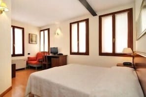 Una camera d'albergo con un letto, una scrivania con una sedia, una televisione e ampie finestre.
