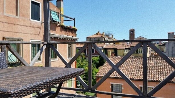 Ein Balkon mit Metallgeländer, der auf ein Wohngebiet mit Terrakotta-Dachziegeln und einem strahlend blauen Himmel blickt.