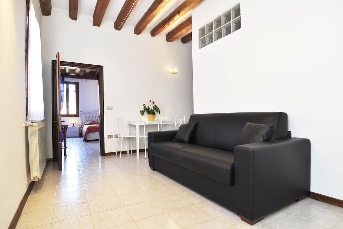 A living room with a black sofa, a small table with a potted plant, and a white chair. There is a doorway leading to another room with a bed and a window.