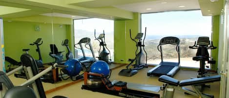 A gym with various exercise equipment, including treadmills and stationary bikes, and a view of the outdoors through large windows.