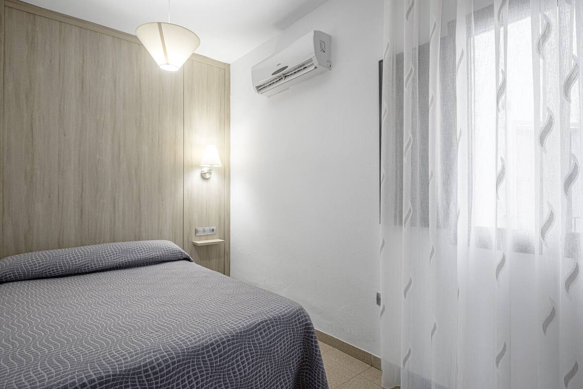 Quarto de hotel com cama, ar condicionado fixado na parede, abajur e cortinas leves.