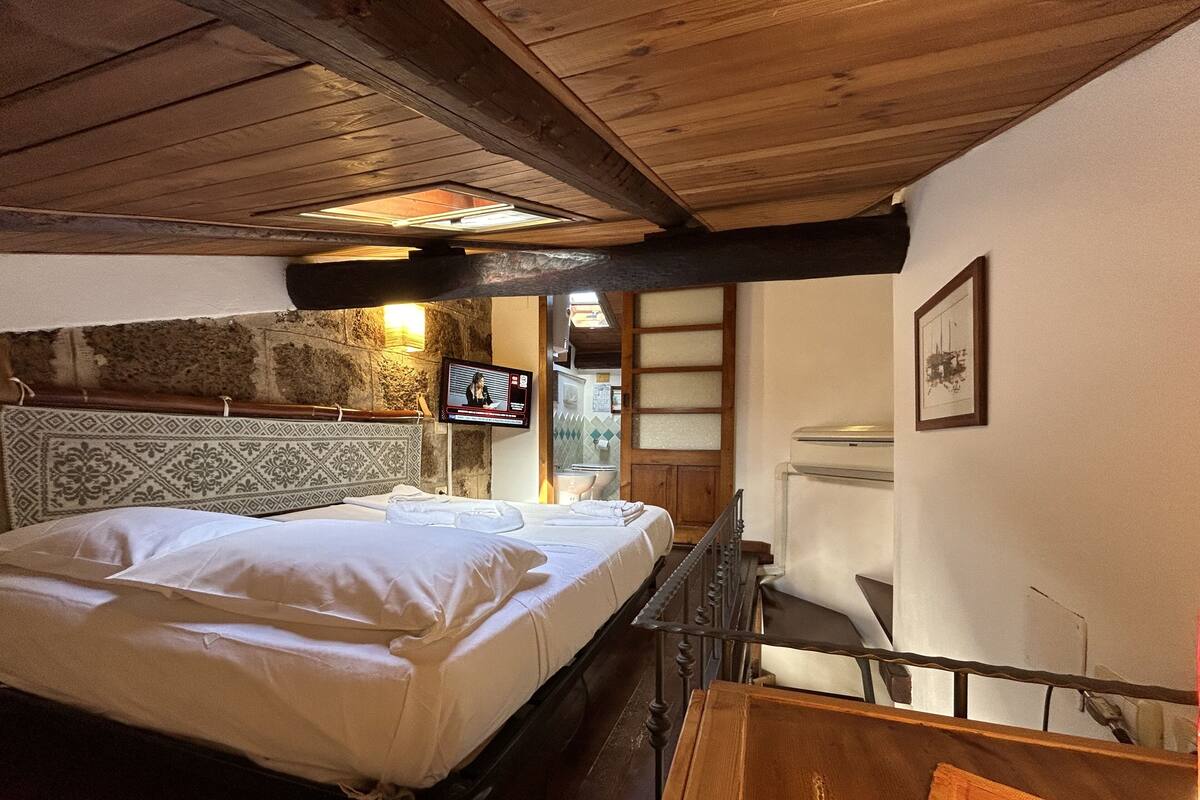 A bedroom with a wooden ceiling, a bed with white linens, a television on the wall, and a stone wall.