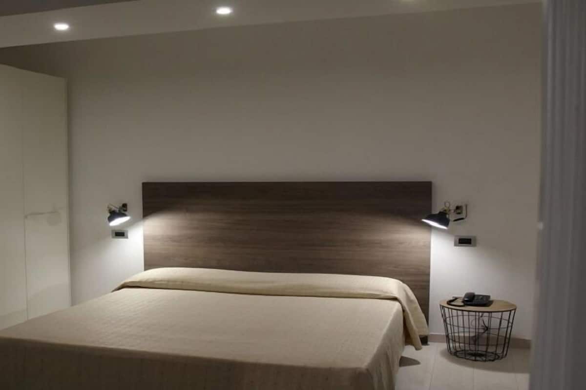 A modern bedroom with a wooden headboard, a bed with a beige bedspread, two wall-mounted reading lamps, and a small round side table with a phone.