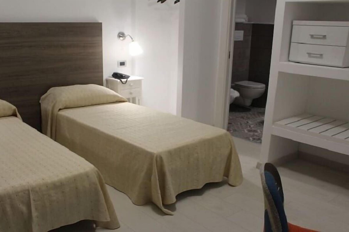 A hotel room with two single beds, a bedside table with a lamp, a wardrobe, and a bathroom visible through an open door.