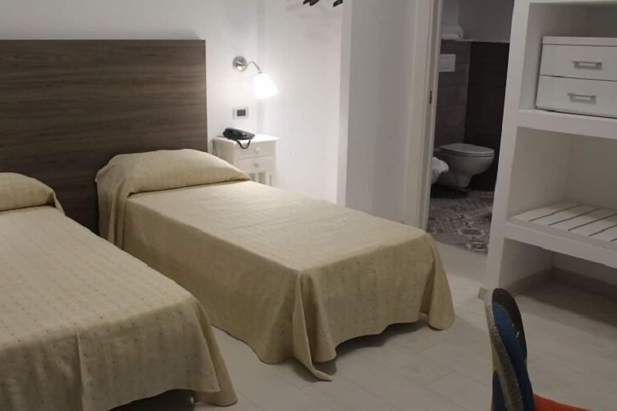 A bedroom with two single beds, a nightstand with a lamp, a wardrobe, and a bathroom visible through an open door.