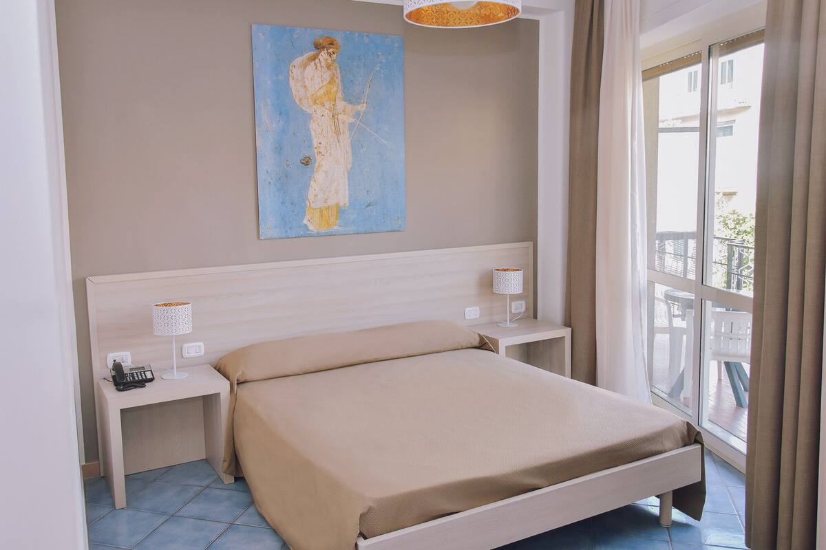 A bedroom with a bed, bedside tables, a lamp, a painting, and a window with curtains.