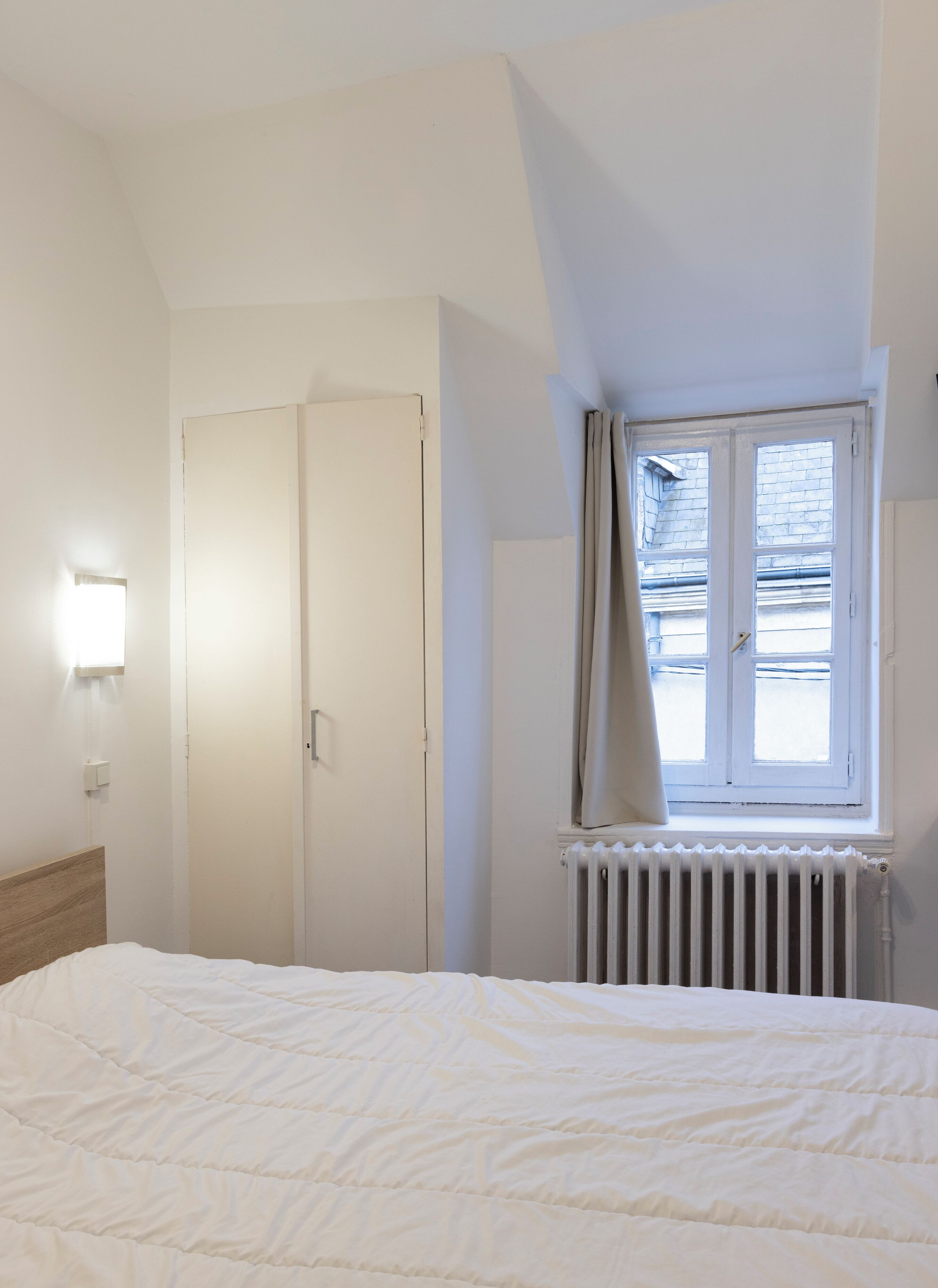 Een slaapkamer met een bed, een radiator, een raam met gordijnen, een deur en een wandlamp.
