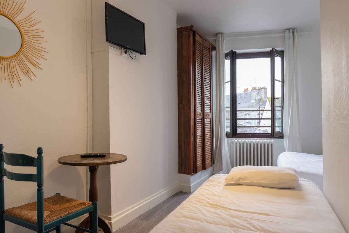 A hotel room with a bed, a chair, a small table, a TV, a window with shutters, and a view of the city.
