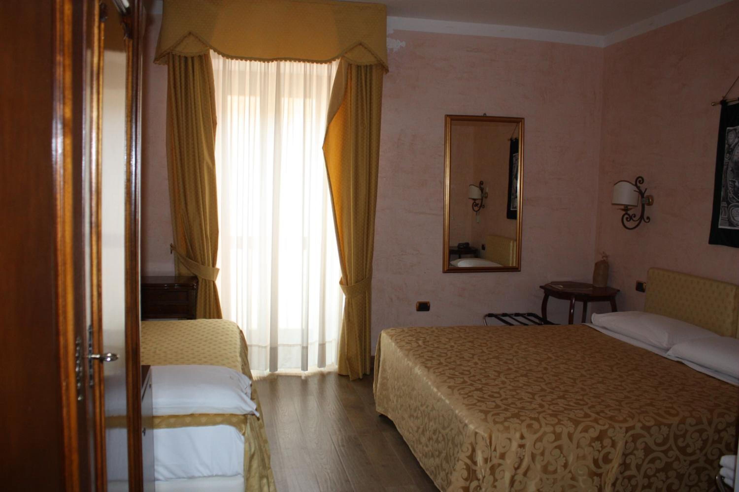 Ein Hotelzimmer mit Bett, Stuhl, kleinem Tisch, Spiegel und einem Fenster mit Vorhängen.