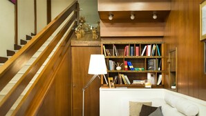 A staircase with wooden railings, a white sofa with cushions, a floor lamp, and a bookshelf with various books and decorative items. - Hotel Orvieto (Orvieto)