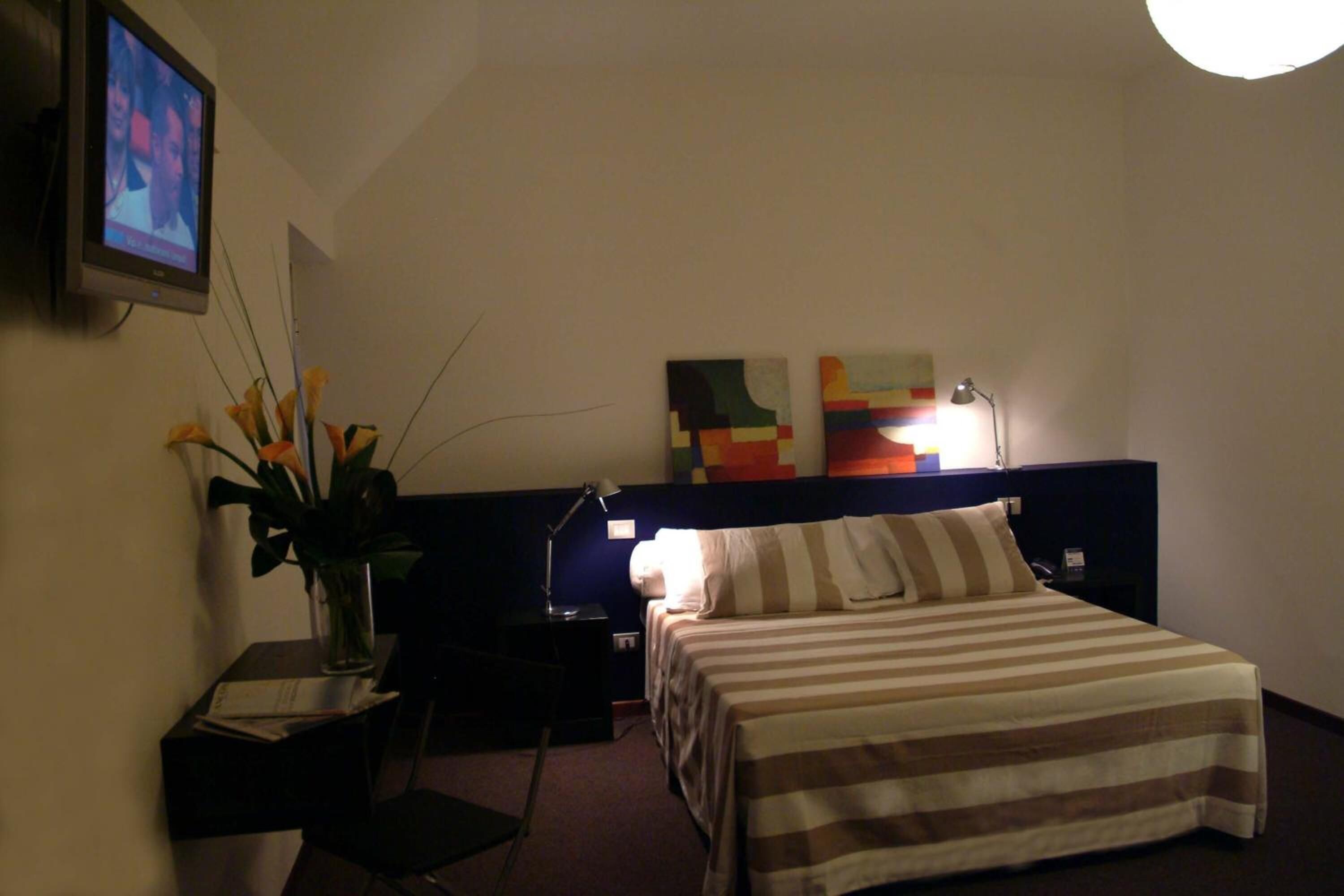 A hotel room with a bed, a nightstand, a lamp, a TV on the wall, and a vase with flowers.