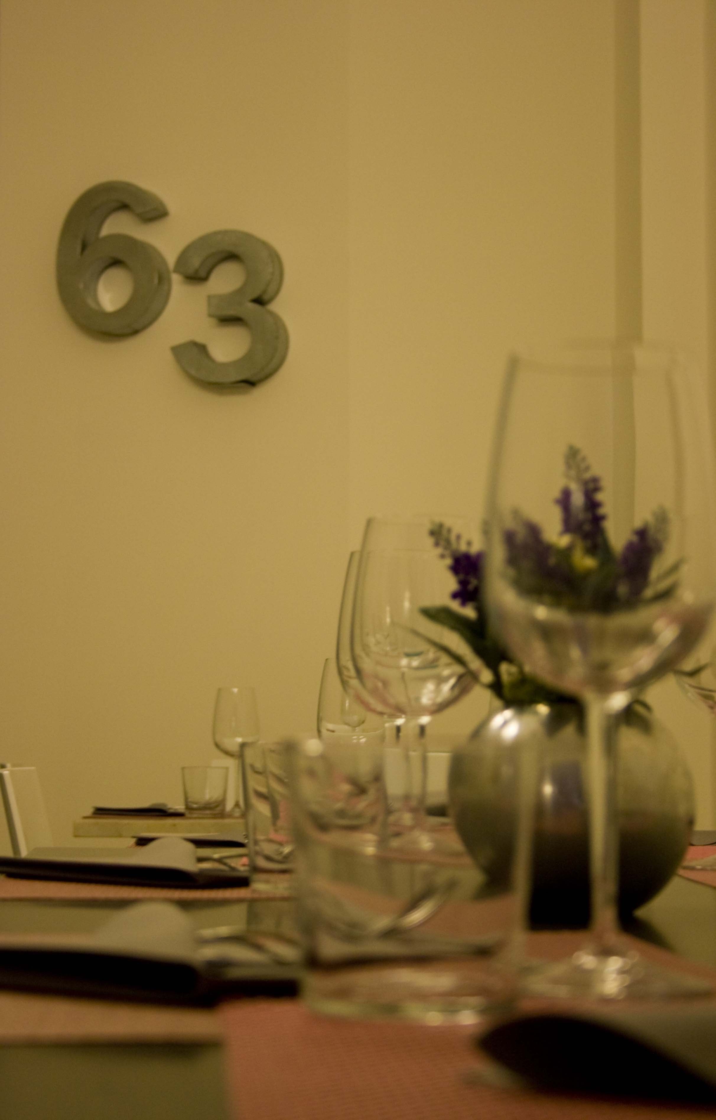 A dining table with wine glasses and a vase with flowers. A number '63' is visible on the wall.