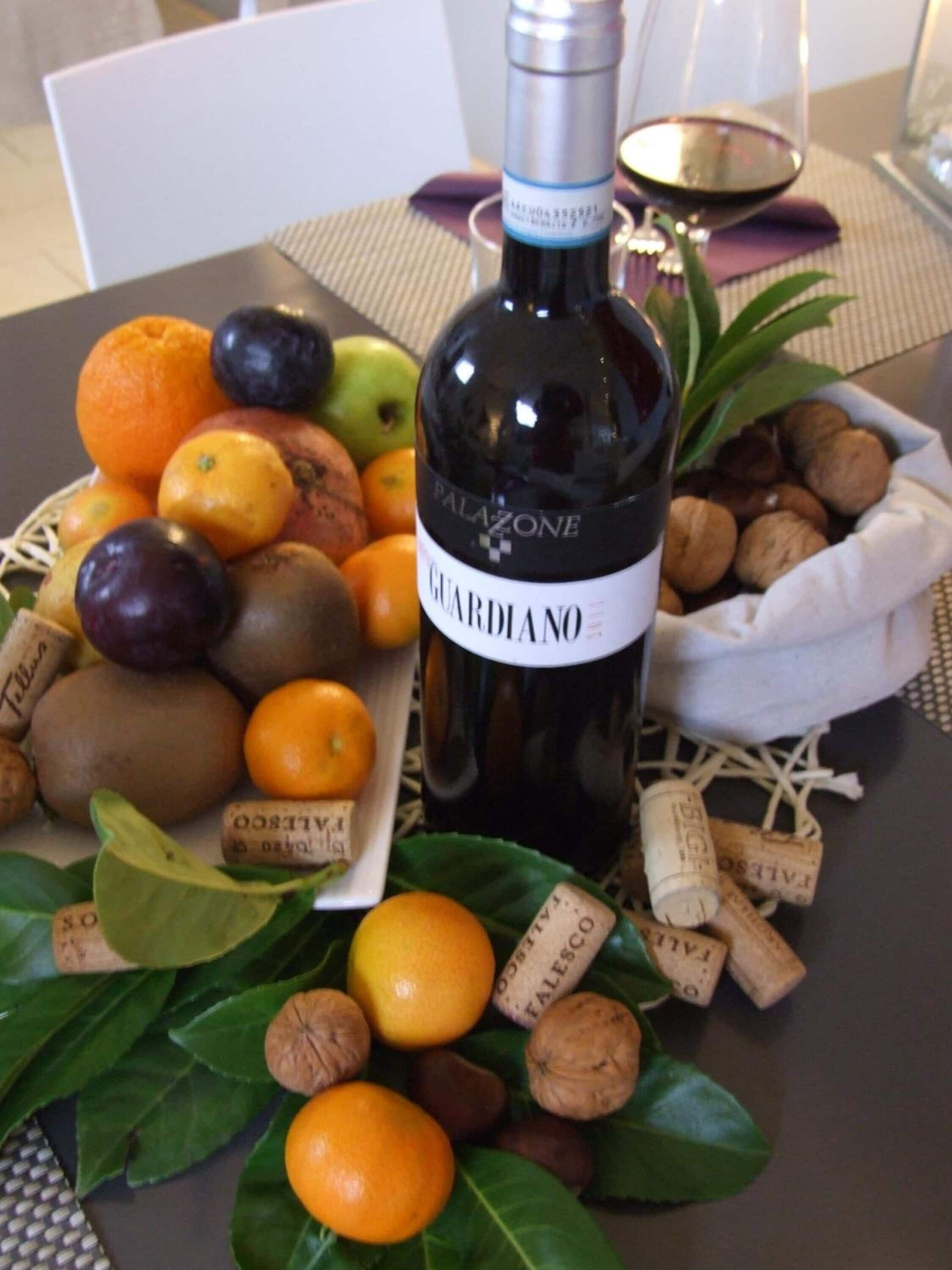 Een fles wijn met de naam 'Palazzo' op een tafel, samen met een fruitmand, noten en wijnkurken.