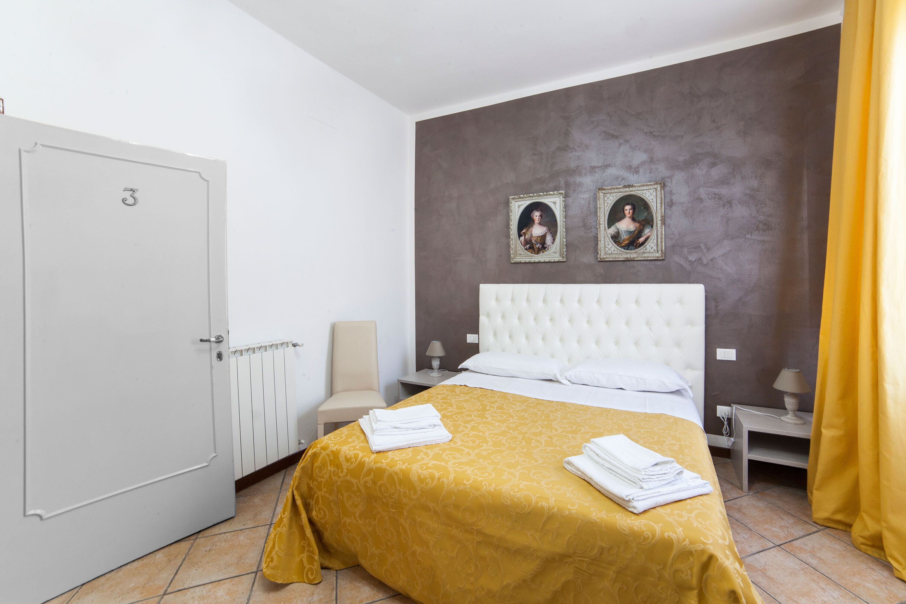 Une chambre d’hôtel avec un lit, une table de chevet, une chaise, une porte et deux tableaux encadrés au mur.