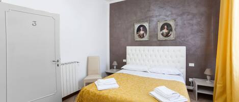 Une chambre d’hôtel avec un lit, une table de chevet, une chaise, une porte et deux tableaux encadrés au mur.