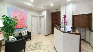 Uma área de recepção com balcão, um espaço de espera com sofá e cadeiras, um grande quadro abstrato e um vaso com planta.