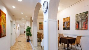 Um corredor com piso ladrilhado, um relógio, obras de arte emolduradas e uma área de jantar com cadeiras e uma mesa.