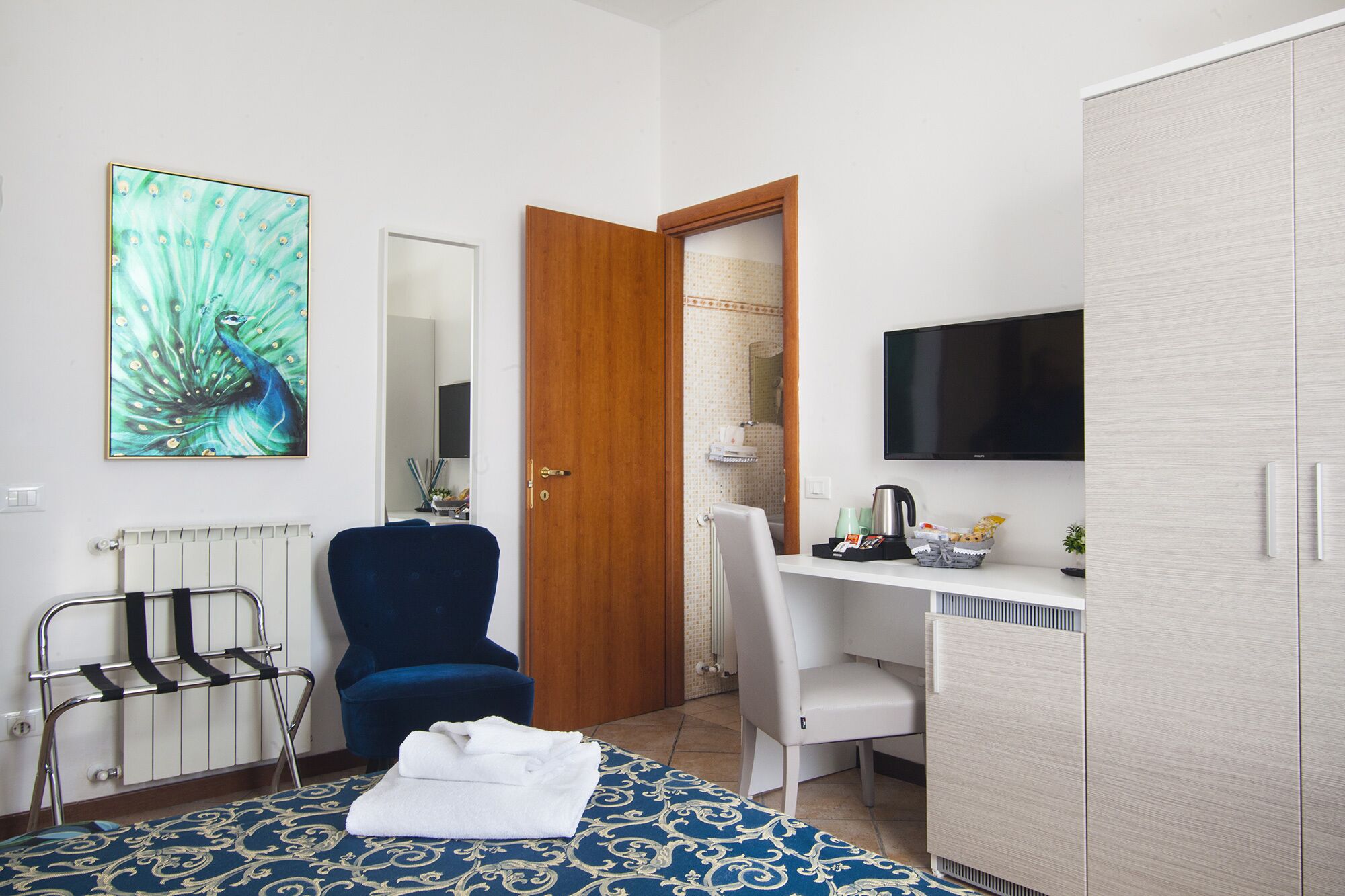 A hotel room with a bed, a desk, a chair, a wardrobe, a radiator, a mirror, and a television.