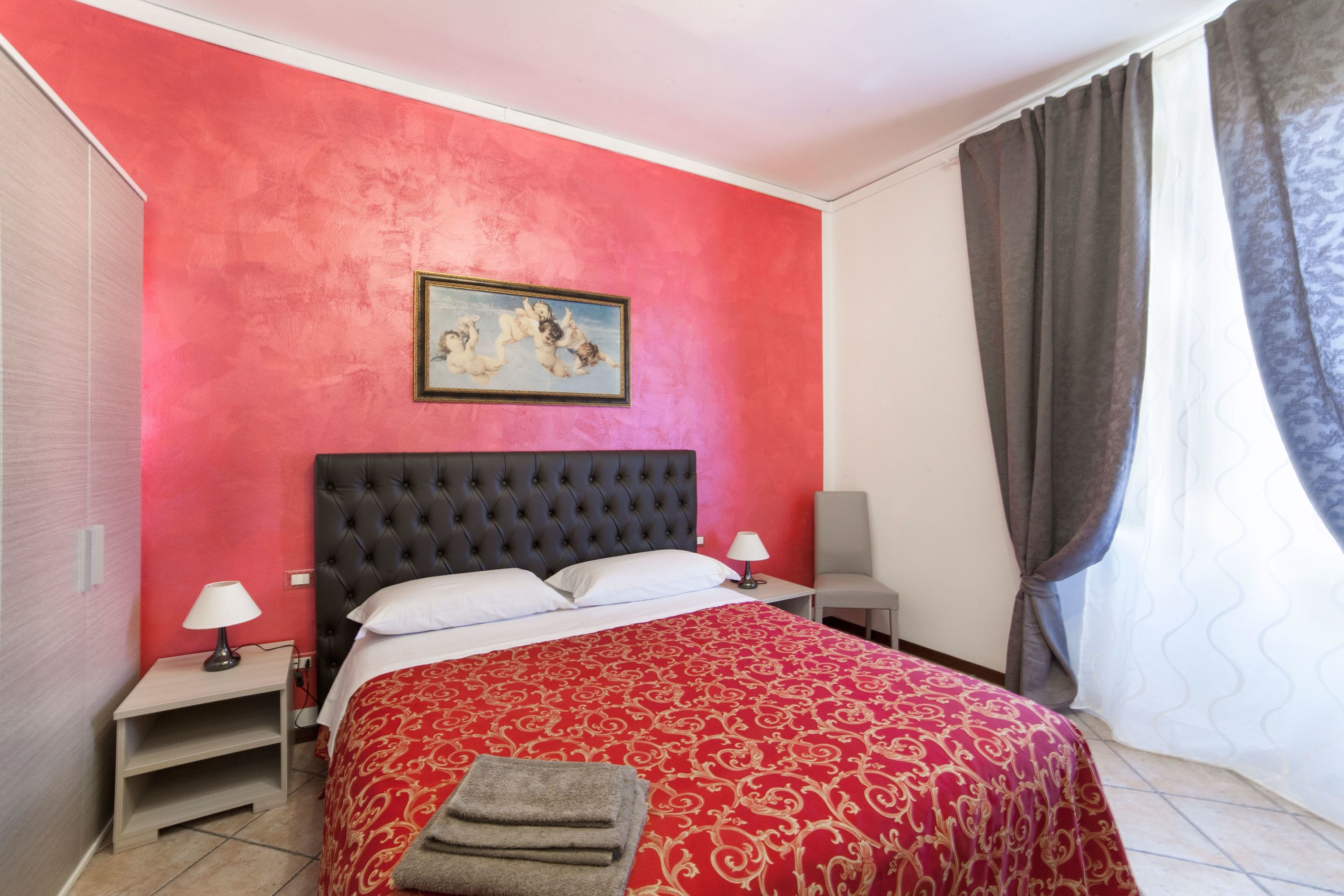 A hotel room with a red wall, a tufted headboard, a bed with a red and gold patterned bedspread, a white pillow, a small bedside table with a lamp, and a chair.