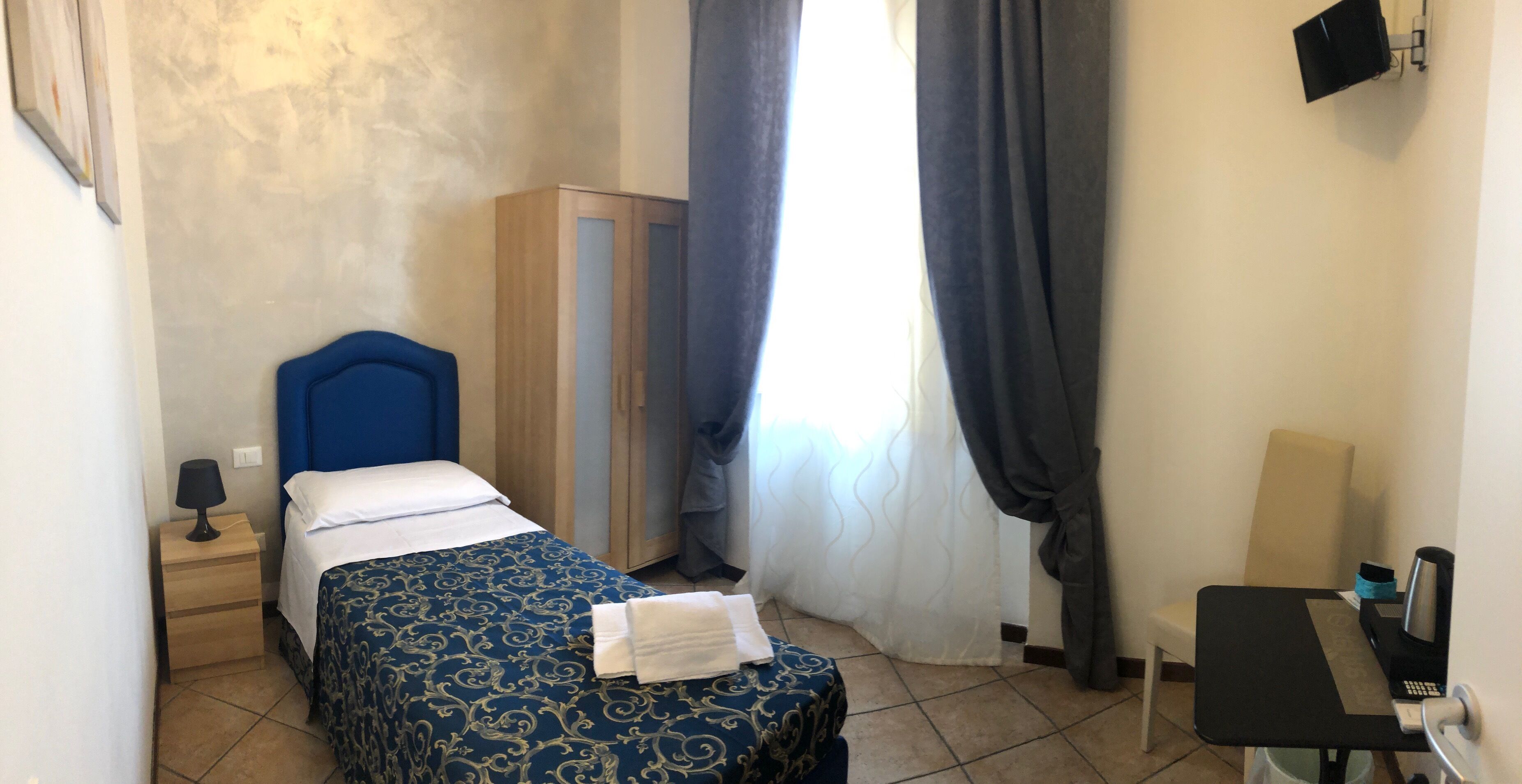 Une chambre d’hôtel comprenant un lit, une table de chevet, un bureau, une armoire et une fenêtre avec des rideaux.