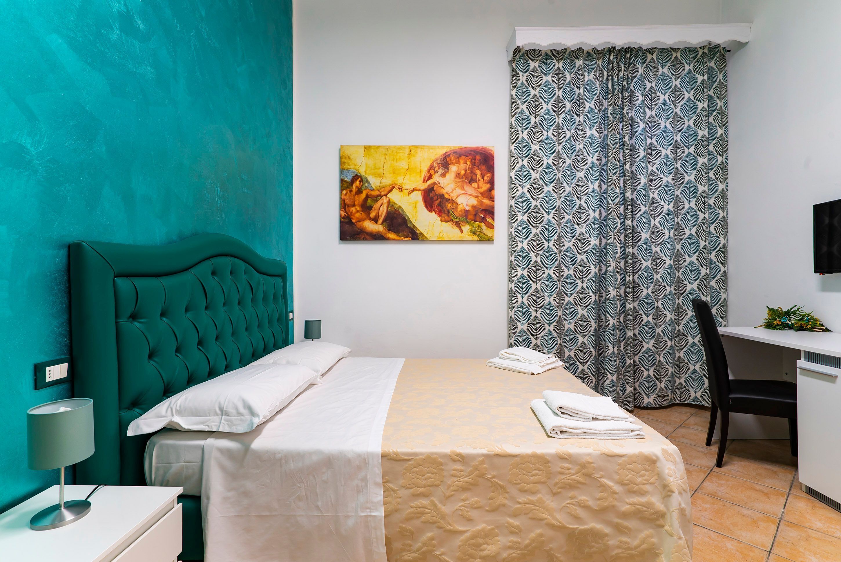 Une chambre d’hôtel avec un mur d’accent de couleur turquoise, un lit recouvert d’une courtepointe blanche et beige, un bureau avec une chaise et un rideau à motifs.