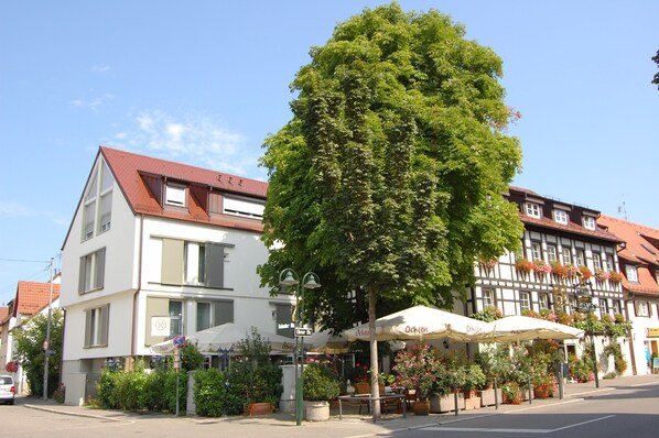 Une vue de rue avec un trottoir bordé d’arbres, un café doté d’une terrasse, et une architecture européenne traditionnelle.