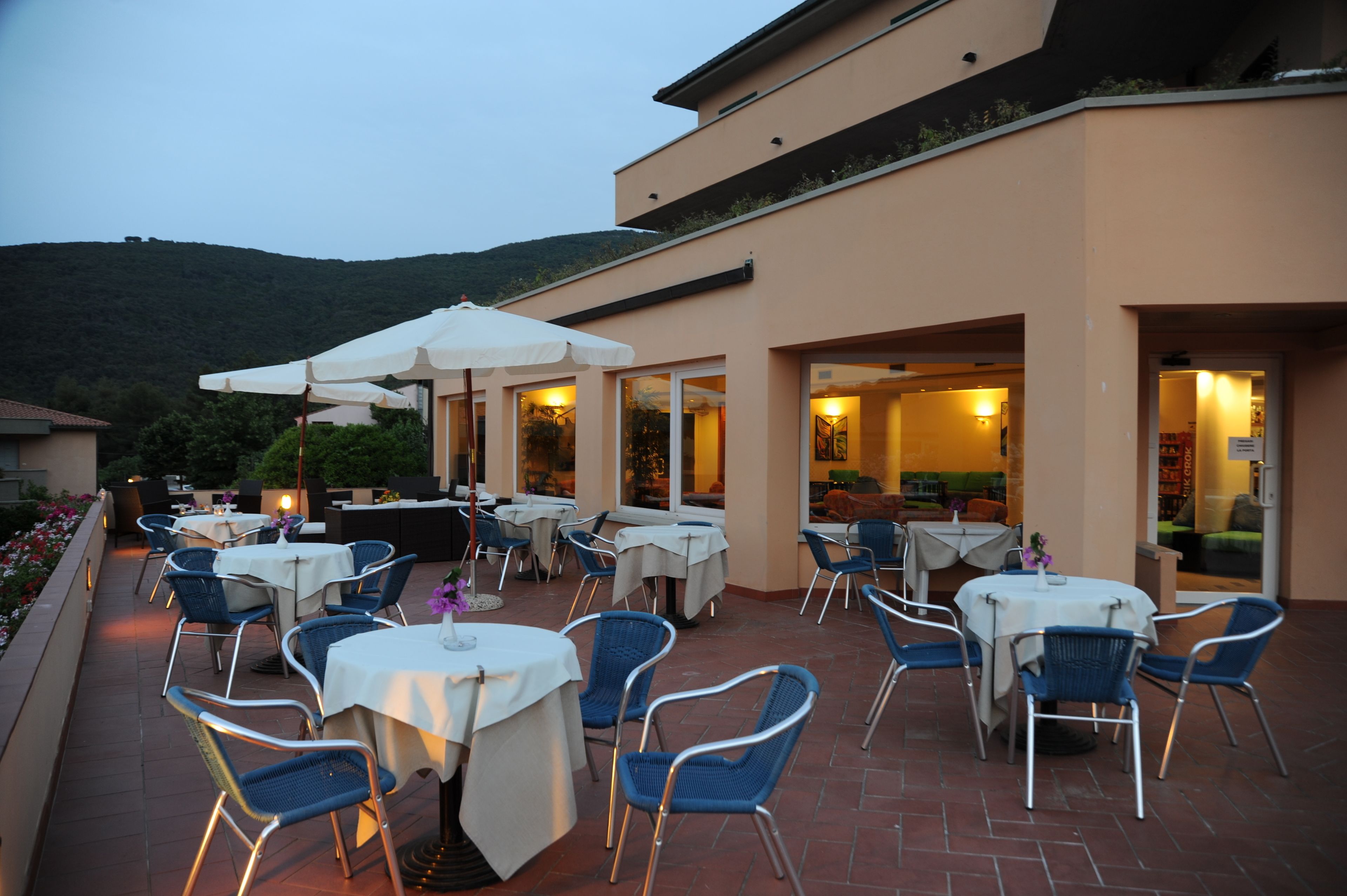 Une terrasse extérieure avec des tables et des chaises, un bâtiment doté de grandes fenêtres et, en arrière-plan, une colline.