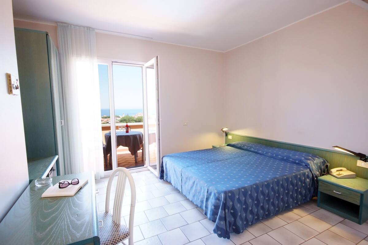 A hotel room with a bed, a desk, a chair, a nightstand, and a view of the sea through a sliding door.