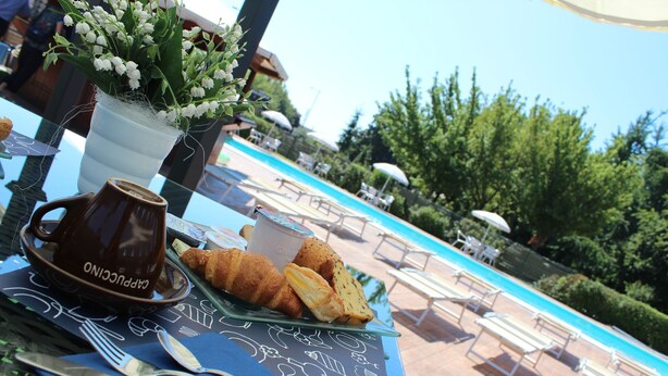 Una mesa con una taza de café, un croissant y un plato de comida, con vista a un área de piscina con sillas de descanso y sombrillas.