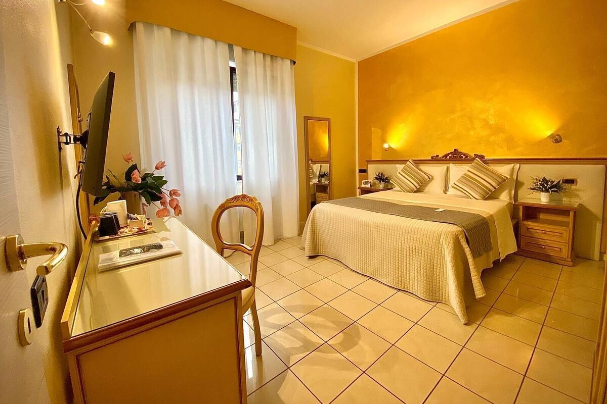 A hotel room with a bed, bedside tables, a desk with a chair, a mirror, and a window with curtains.