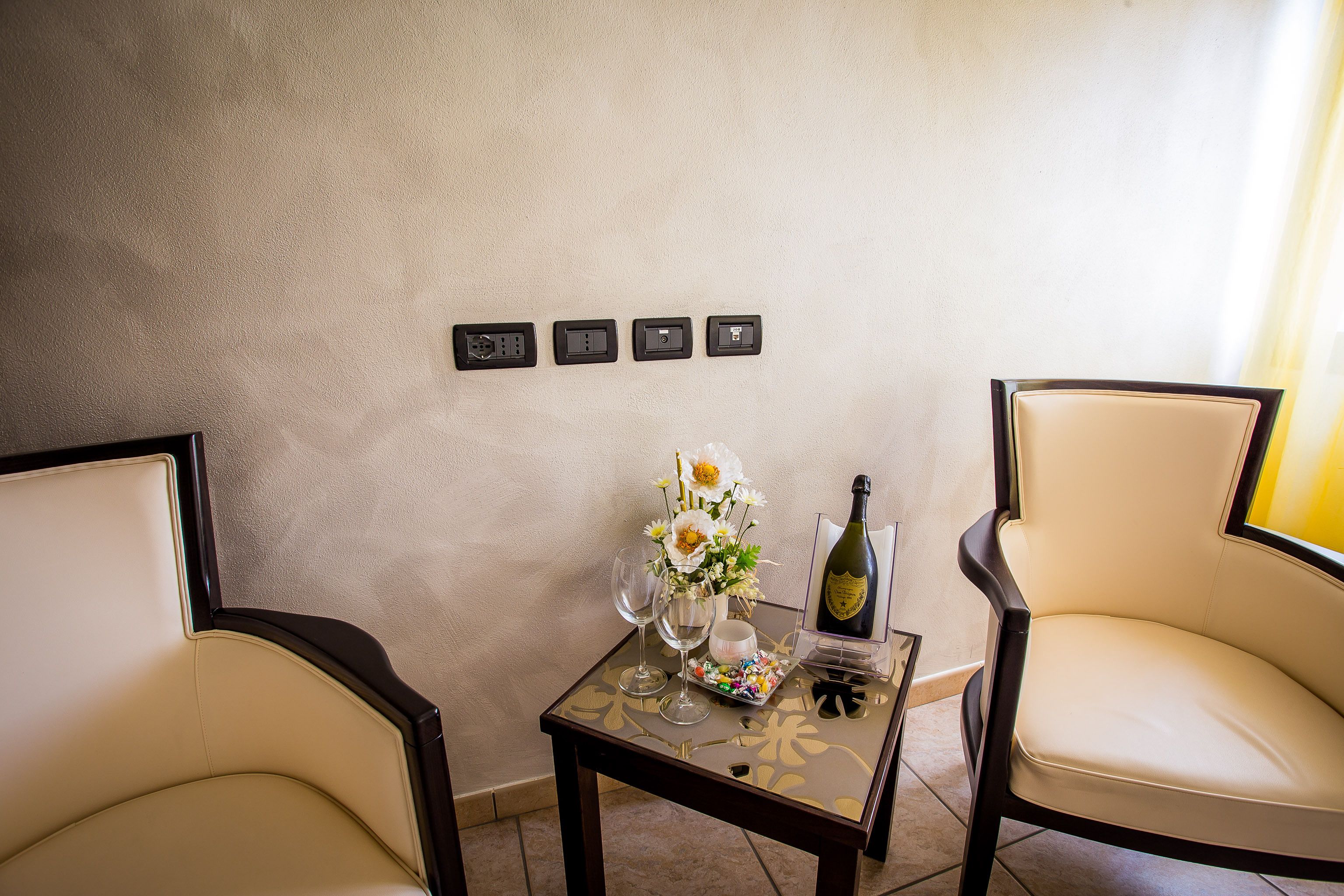Ein Zimmer mit zwei Stühlen, einem kleinen Tisch mit Blumenvase, zwei Weingläsern, einer Weinflasche und drei Steckdosen an der Wand.