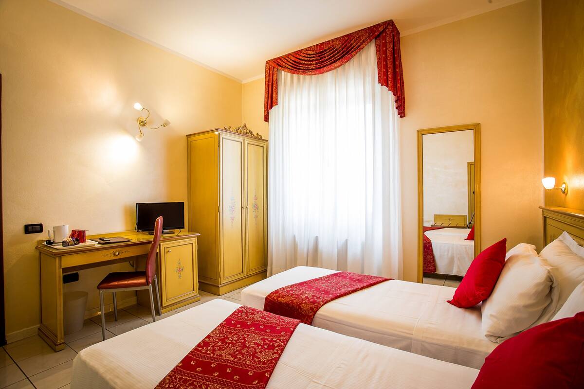 A hotel room with two beds, a desk, a chair, a wardrobe, and a large window with curtains.