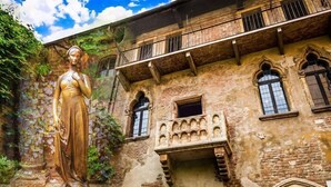 Un edificio histórico con una estatua frente a él, que presenta ventanas en arco y un balcón con barandilla de hierro forjado.